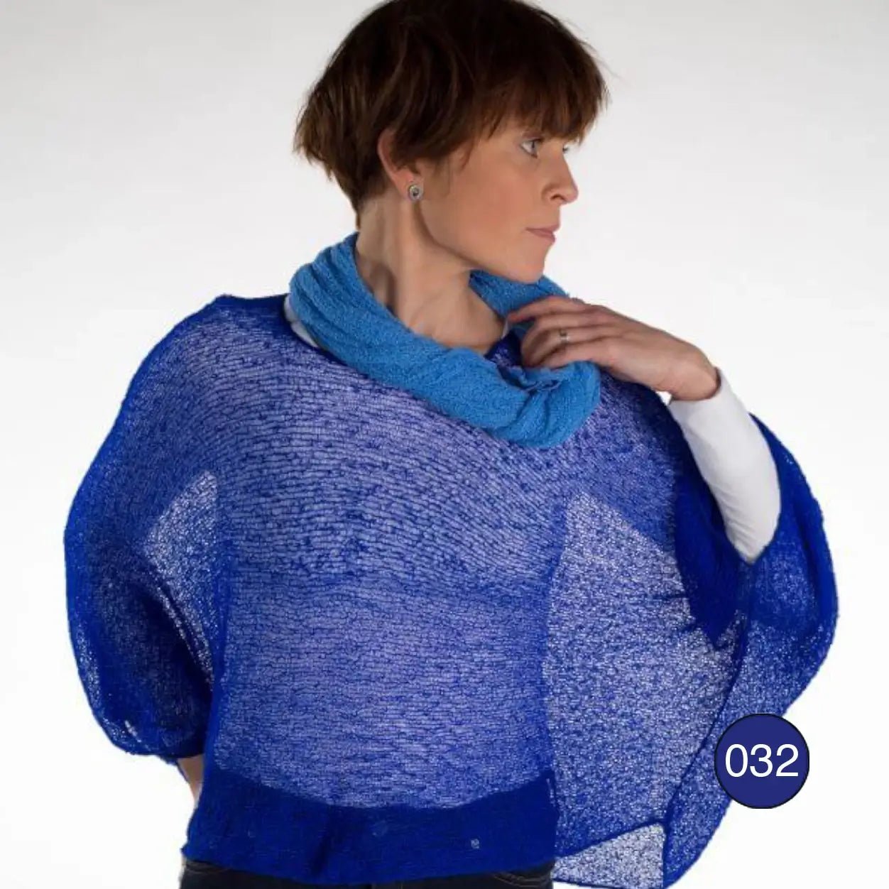 Korthårig kvinna i en klarblå Lebar poncho med ärm; det tunna finstickade garnet i klar kobolt faller löst och draperat över axlar och armar i ett luftigt fall. Ulla Jacobsson Kläder