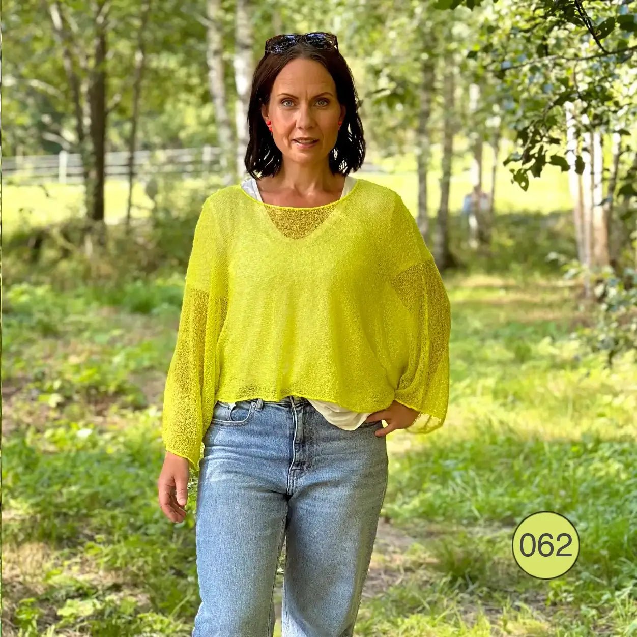 Mörkhårig kvinna i en limegrön Lebar poncho över jeans i grönskan; det finstickade garnet i frisk gulgrön ton faller löst över en ljus topp och draperar mjukt över axlarna. Ulla Jacobsson Kläder