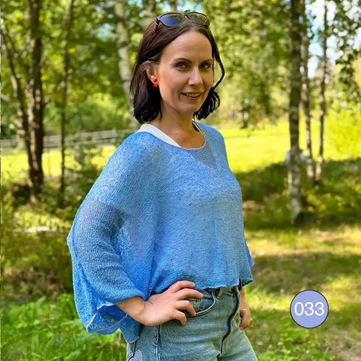 Mörkhårig kvinna med solglasögon i håret och handen i sidan, i en ljusblå Lebar poncho i björkskog; det finstickade garnet faller mjukt över jeans i ett lätt, draperat lager. Ulla Jacobsson Kläder