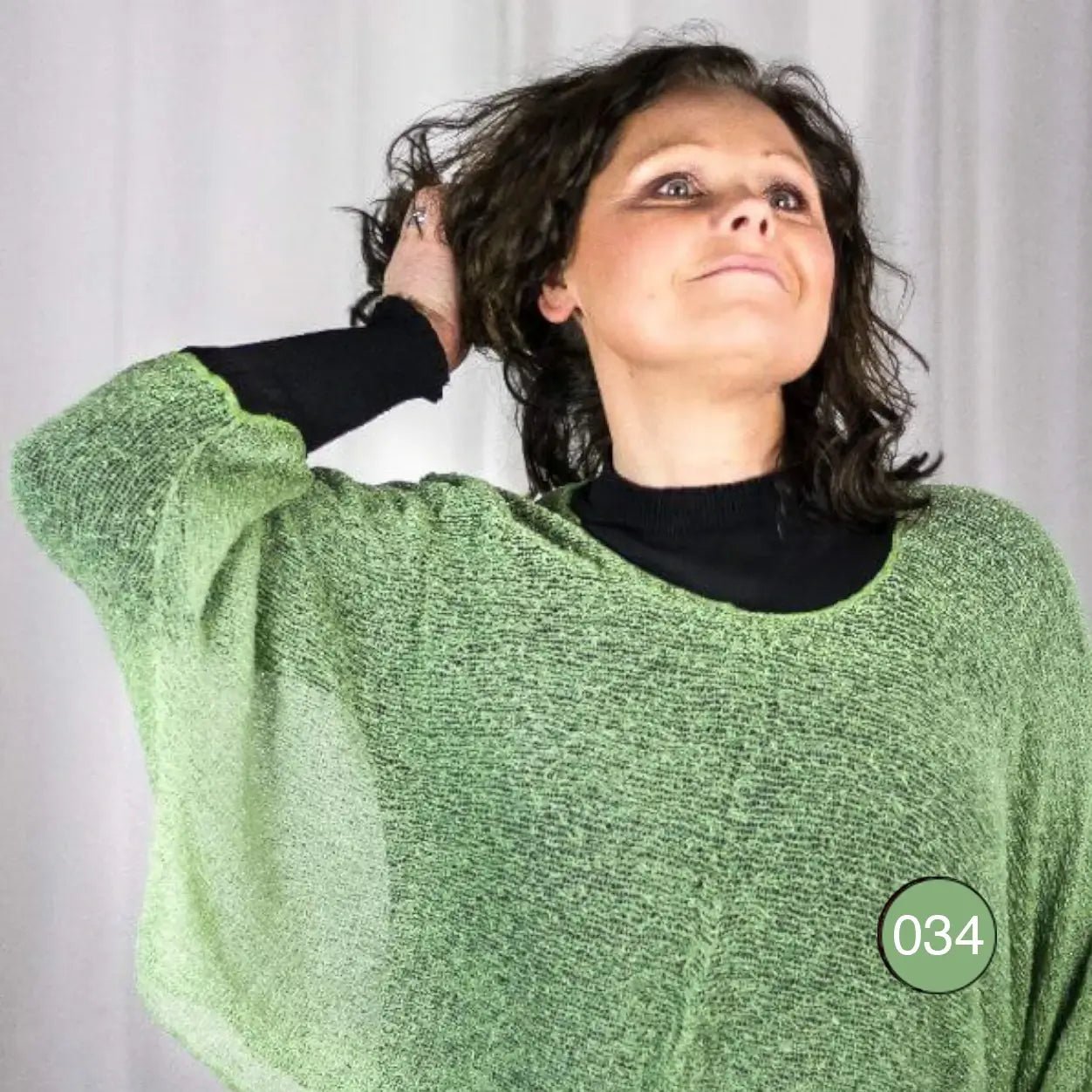 Mörkhårig kvinna lutar huvudet bakåt i en ljusgrön Lebar poncho; det finstickade garnet i frisk grönton draperar löst över en svart underdel och de vida ärmarna faller mjukt. Ulla Jacobsson Kläder