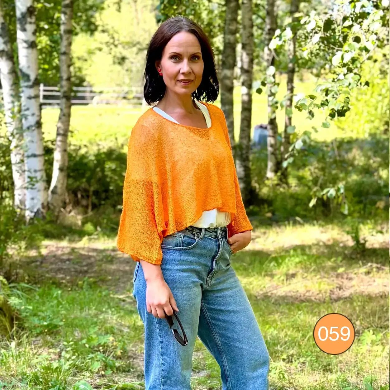 Mörkhårig kvinna i björkskog i en ljusorange Lebar poncho över jeans; det tunna finstickade garnet lyser varmt orange och faller draperat över axlar och armar. Ulla Jacobsson Kläder