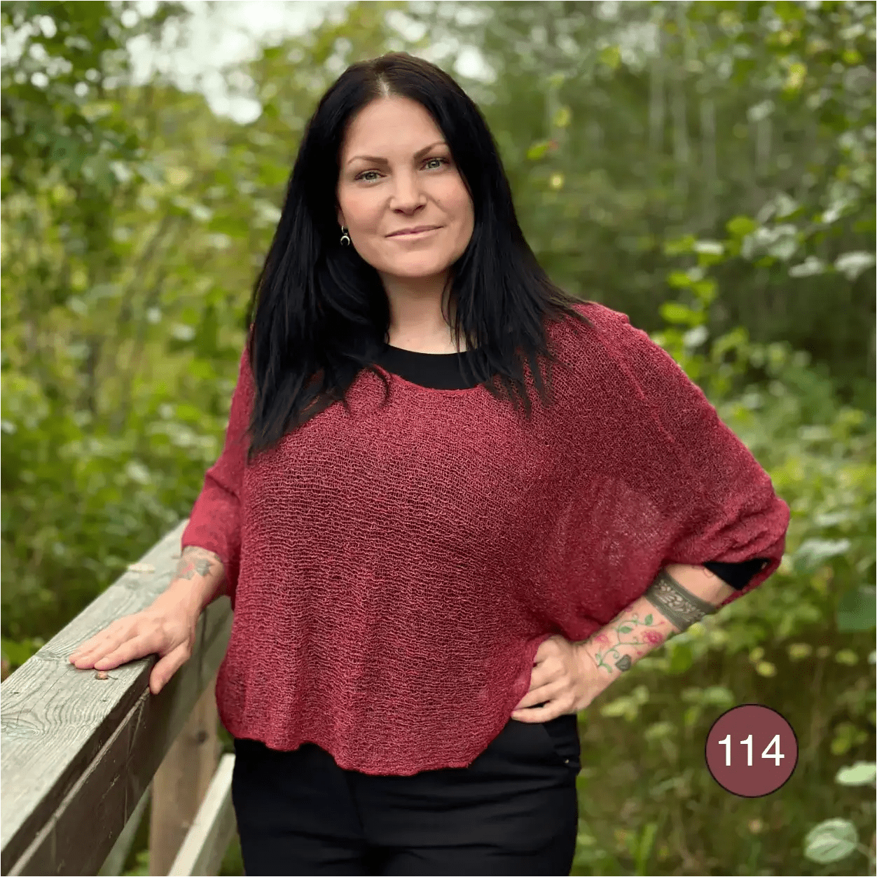 Mörkhårig kvinna vid ett räcke i en Lebar poncho i färgen maroon; den tvåtonade finstickade ponchon med vida ärmar faller mjukt och draperat över svart. Ulla Jacobsson Kläder