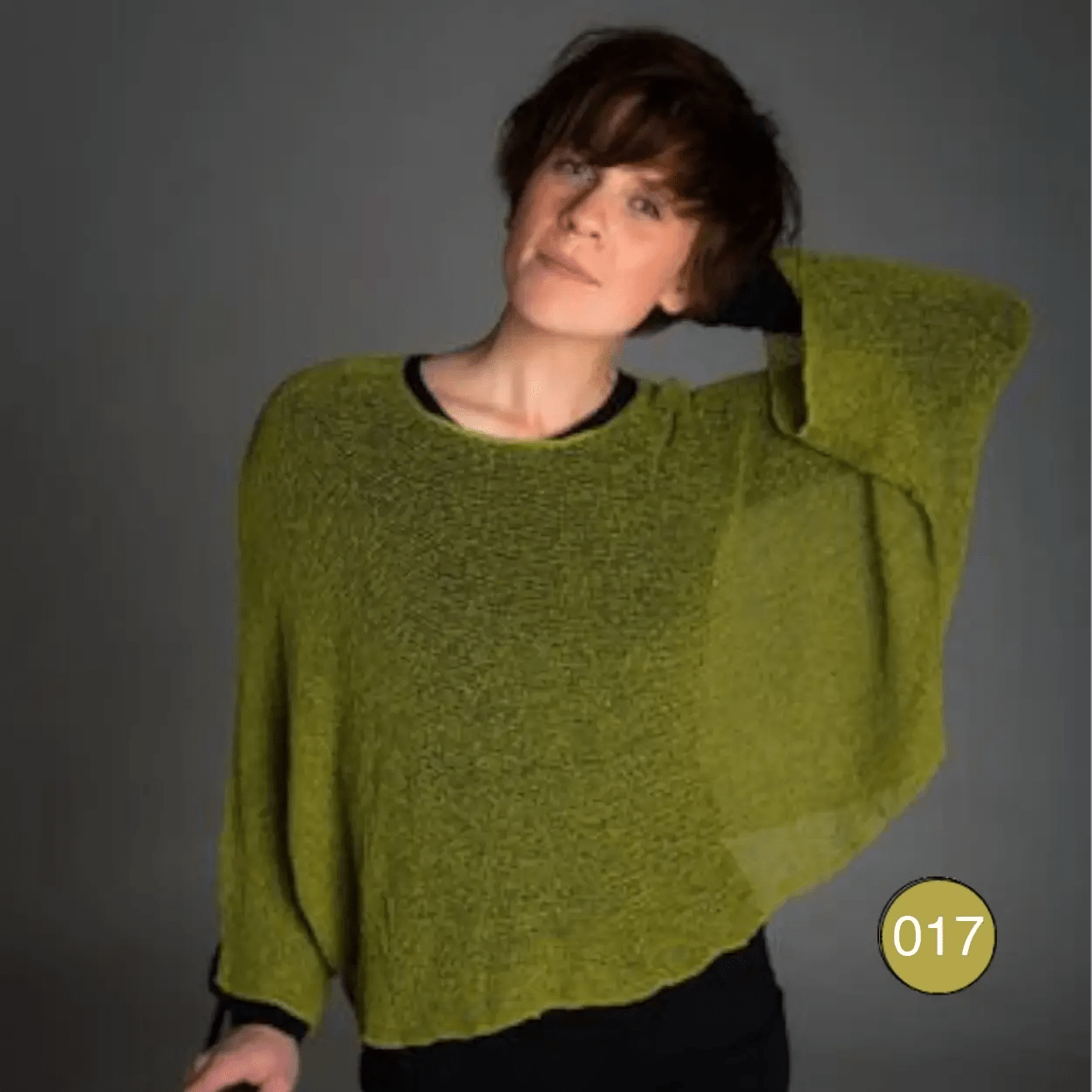 Korthårig kvinna i en olivgrön Lebar poncho med ena armen lyft så den vida ärmen syns; det finstickade garnet faller i djup olivton löst och draperat över en svart underdel. Ulla Jacobsson Kläder