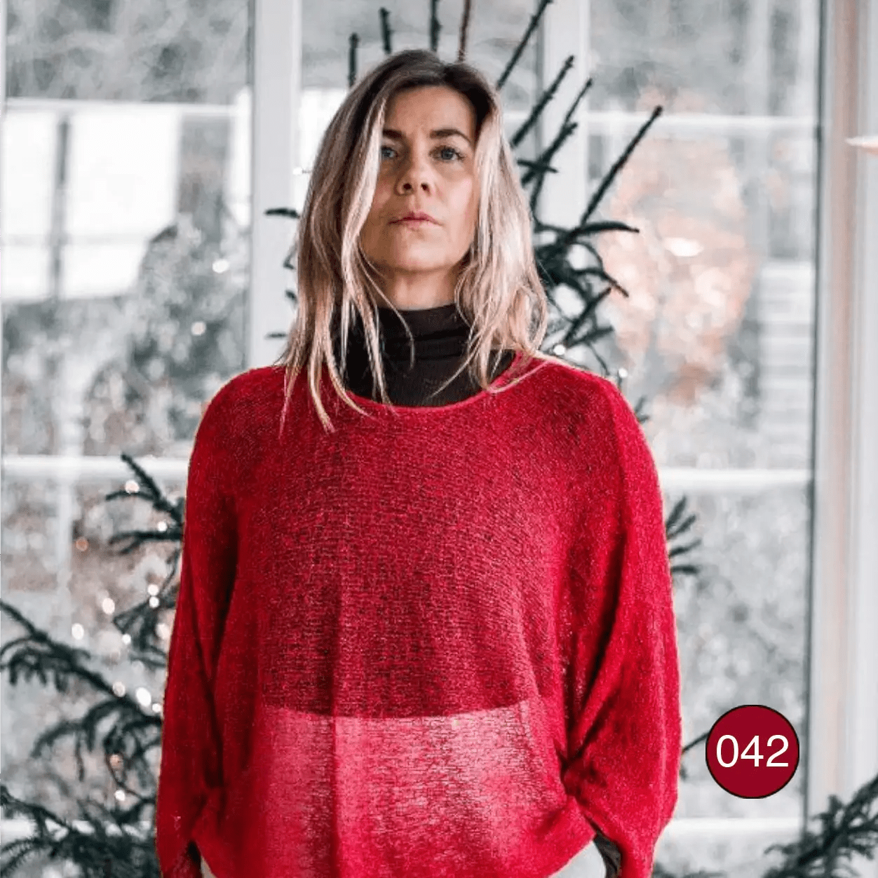 Ljushårig kvinna i en röd Lebar poncho med ärm över en svart polotröja; den finstickade ponchon skiftar i två röda toner och faller mjukt och draperat över axlar och armar. Ulla Jacobsson Kläder