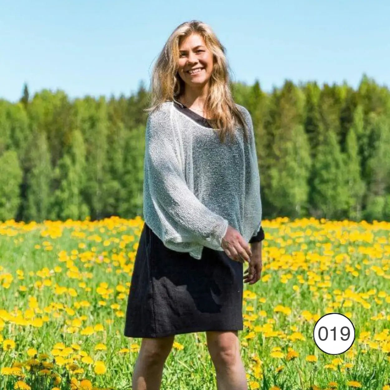 Ljushårig kvinna i en sommaräng full av maskrosor bär en vit Lebar poncho över en mörk kjol; det luftiga finstickade garnet lyser ljust i solen och faller mjukt över axlar och armar. Ulla Jacobsson Kläder