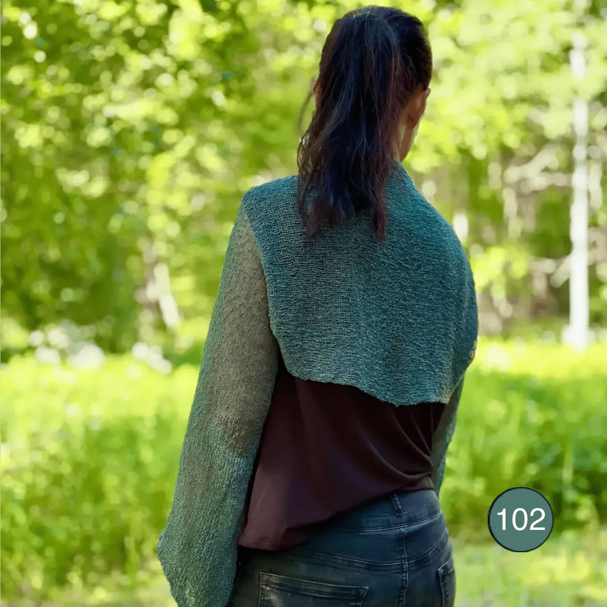 Mörkhårig kvinna fotograferad bakifrån i en buteljgrön Liv bolero ovanpå en mörk topp; den korta finstickade boleron faller luftigt över ryggen i en djup, dämpad grönton. Ulla Jacobsson Kläder