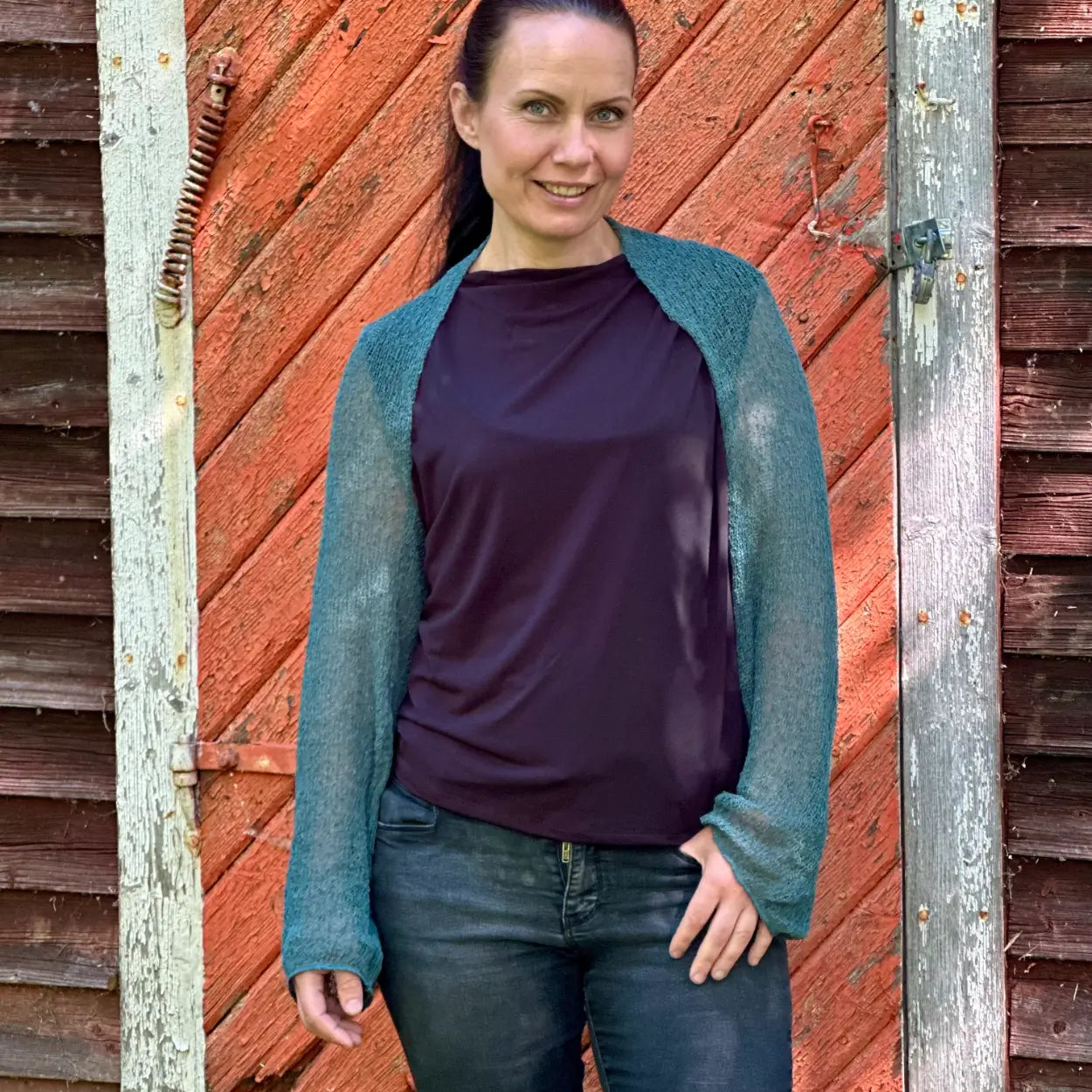 Mörkhårig kvinna mot en tegelvägg i en buteljgrön Liv bolero över en mörk topp och jeans; det finstickade garnet i djup grönton sitter tätt över axlarna och de långa ärmarna. Ulla Jacobsson Kläder