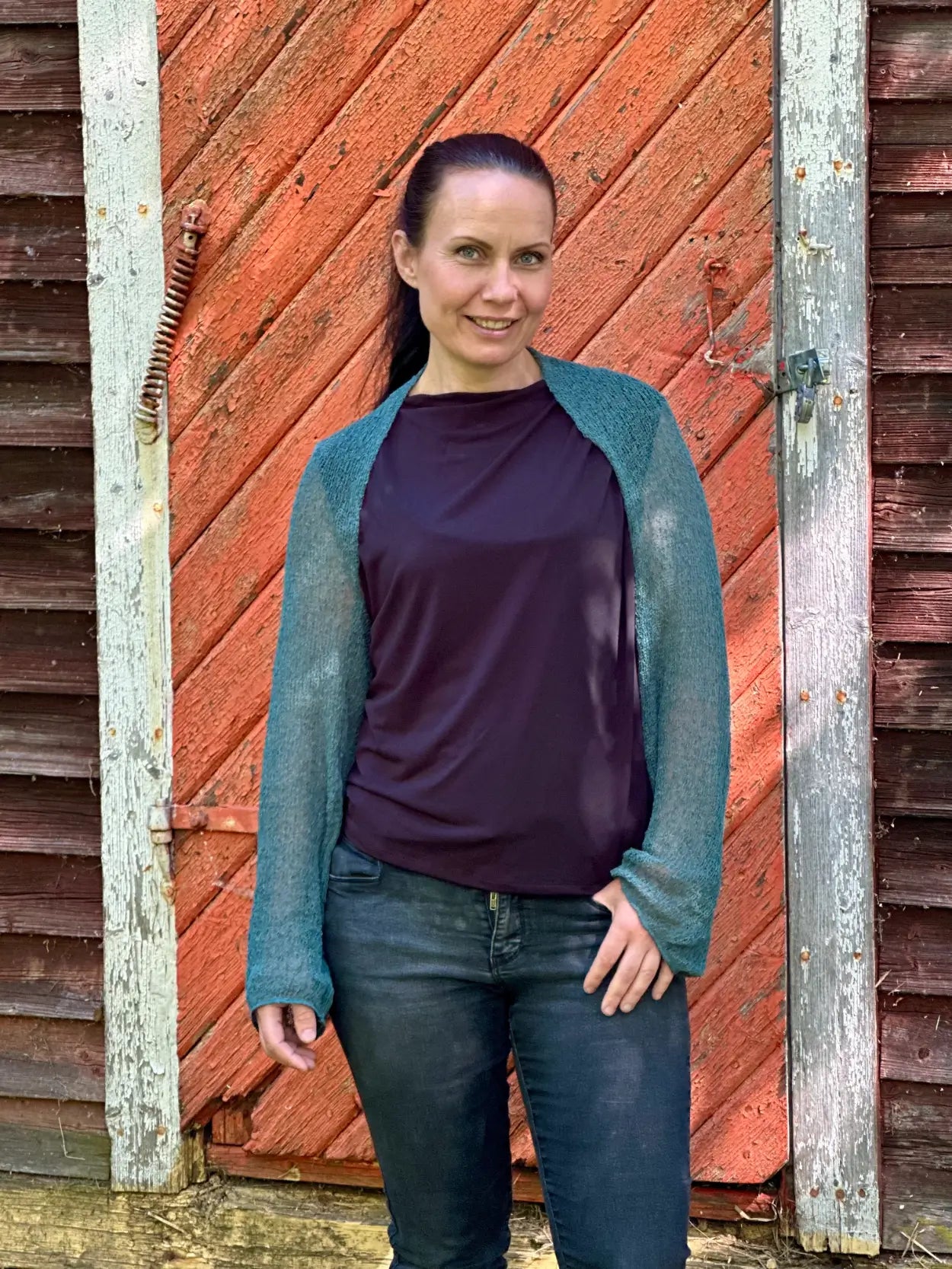 Mörkhårig kvinna mot en tegelvägg i en buteljgrön Liv bolero över en mörk topp och jeans; det finstickade garnet i djup grönton sitter tätt över axlarna och de långa ärmarna. Ulla Jacobsson Kläder