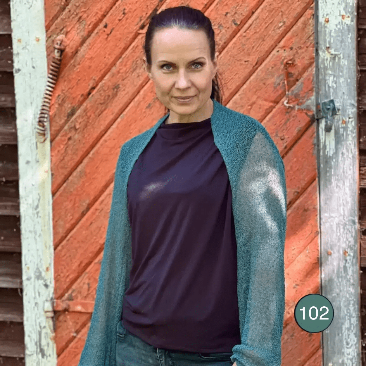 Mörkhårig kvinna mot en röd tegelvägg i en buteljgrön Liv bolero över en mörk topp; det finstickade garnet i djup grönblå ton sitter tätt över axlarna och de långa ärmarna. Ulla Jacobsson Kläder