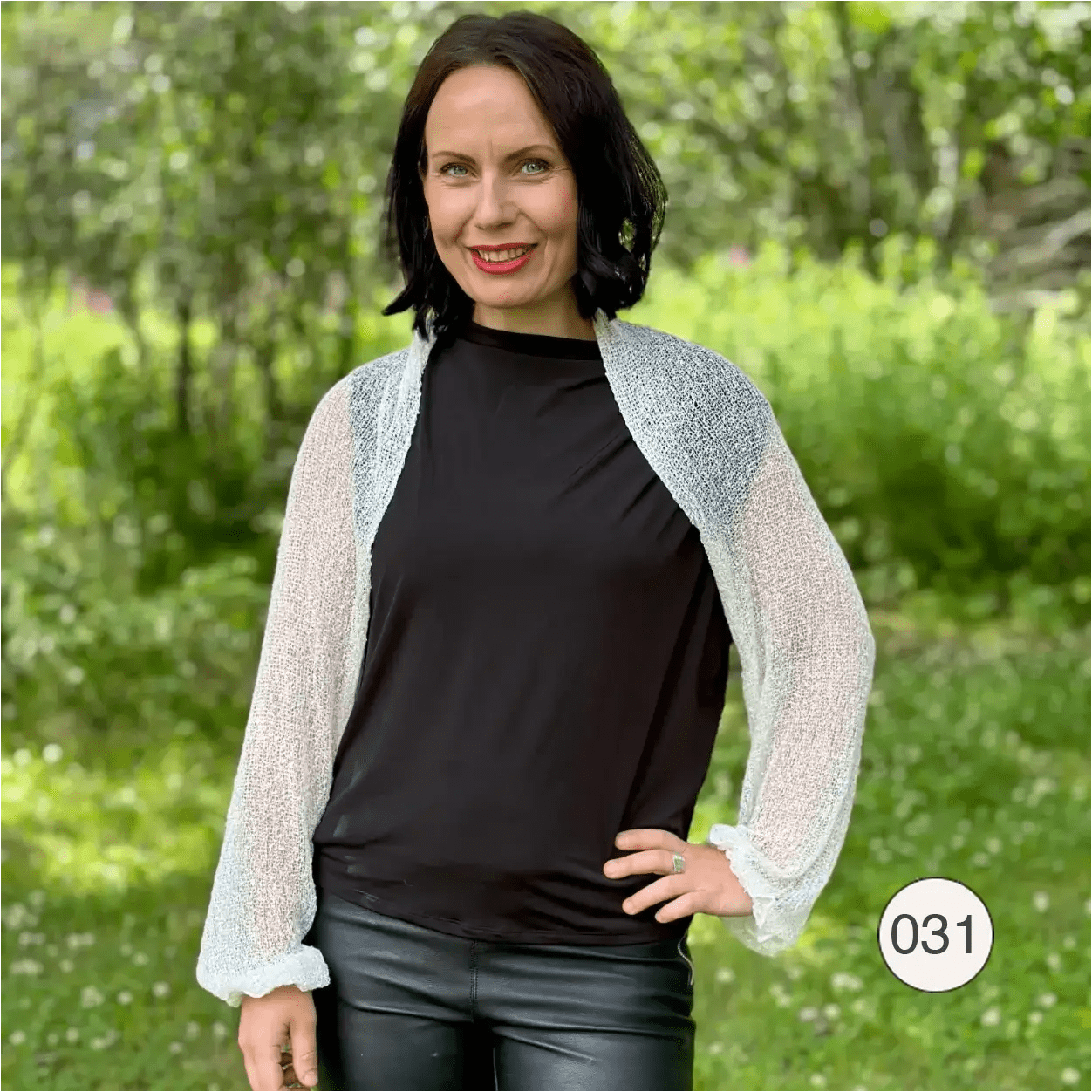 Mörkhårig kvinna med handen i sidan i en offwhite Liv bolero buren över svart topp och skinnbyxor; det tunna, lätta finstickade garnet ger en mjuk, luftig kontrast mot den svarta outfiten. Ulla Jacobsson Kläder