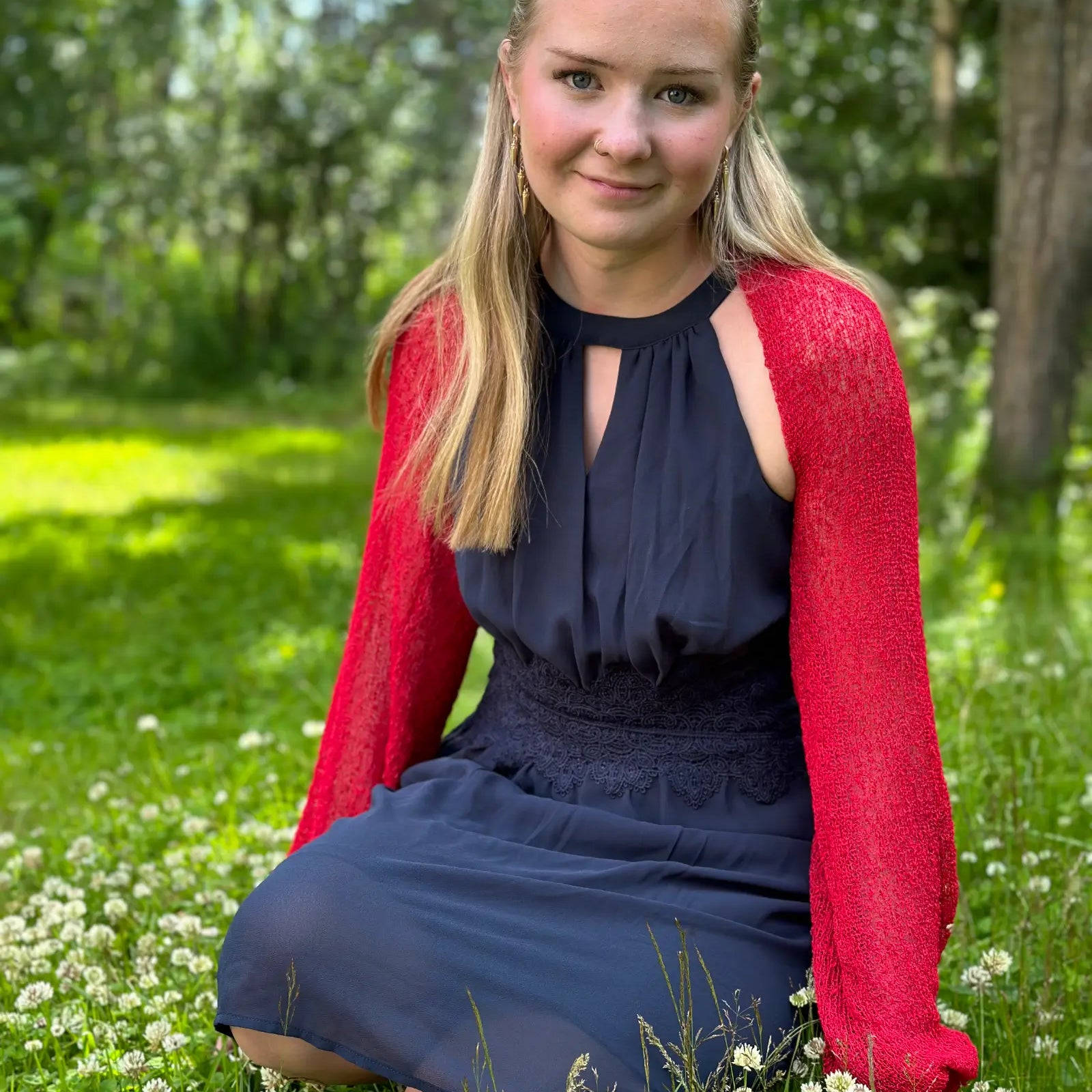 Ljushårig kvinna på en sommaräng i en röd Liv bolero buren över en marinblå klänning; den finstickade boleron med långa ärmar lyser klarrött mot grönskan och värmer axlarna en ljum kväll. Ulla Jacobsson Kläder