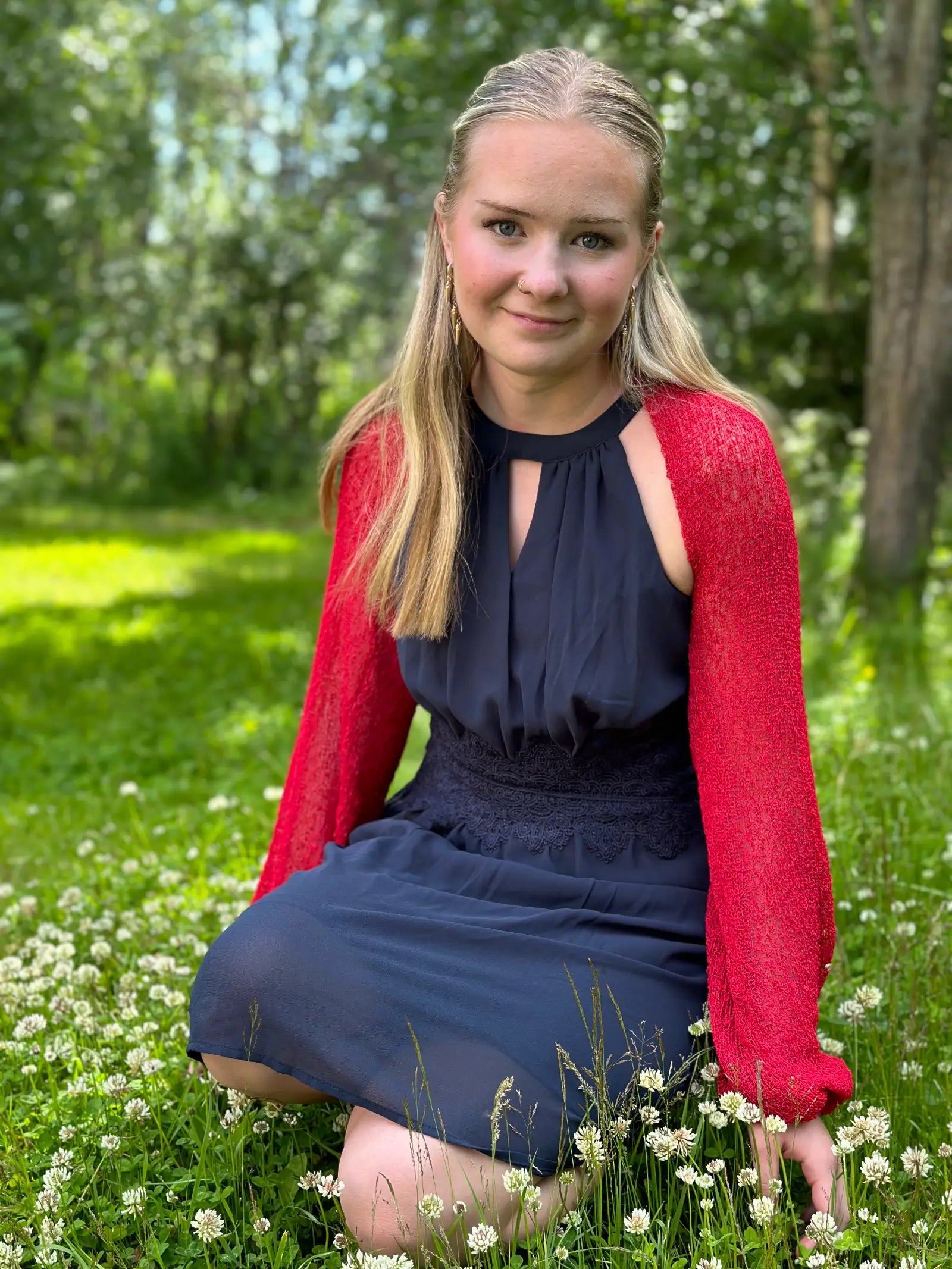 Ljushårig kvinna på en sommaräng i en röd Liv bolero buren över en marinblå klänning; den finstickade boleron med långa ärmar lyser klarrött mot grönskan och värmer axlarna en ljum kväll. Ulla Jacobsson Kläder