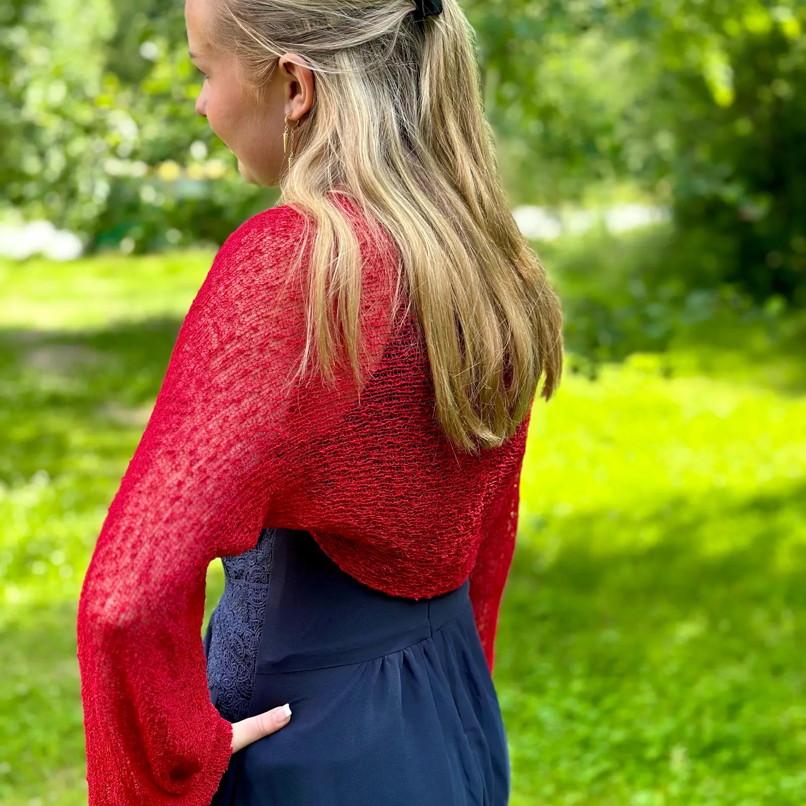 Ljushårig kvinna fotograferad bakifrån i en röd Liv bolero ovanpå en marinblå klänning; den korta finstickade boleron faller över ryggen och de långa ärmarna avslutas med resår vid handlederna. Ulla Jacobsson Kläder