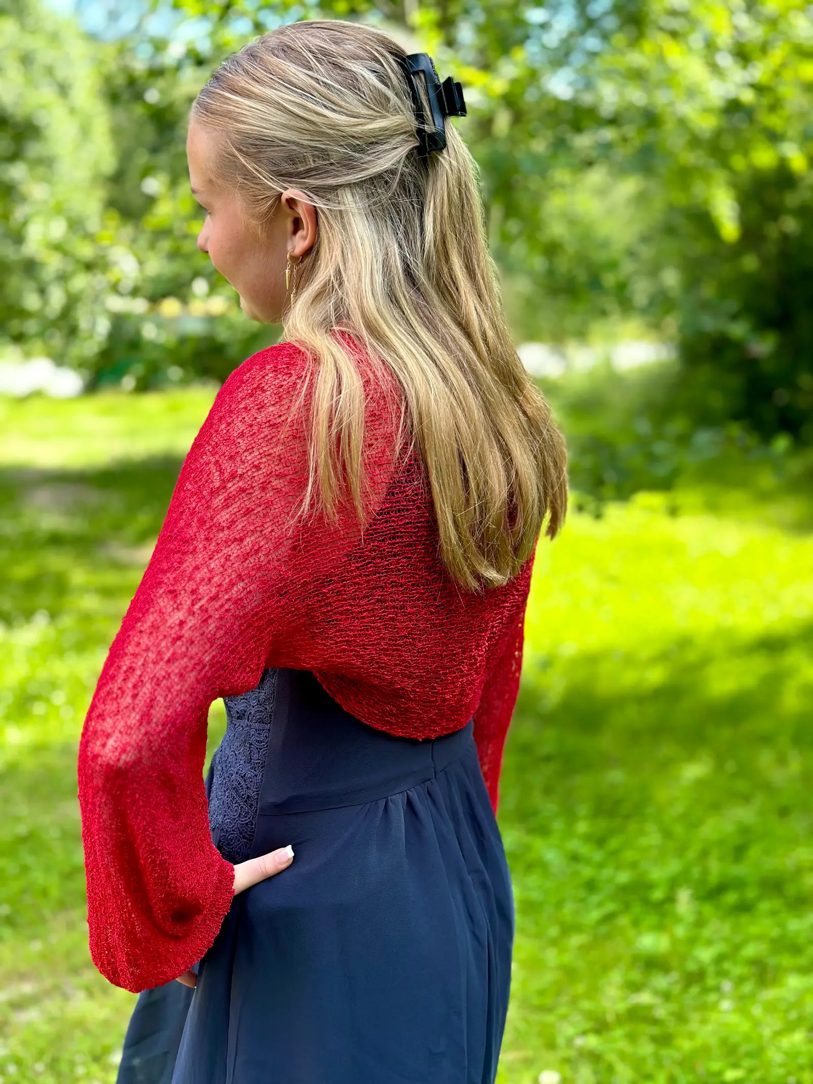 Ljushårig kvinna fotograferad bakifrån i en röd Liv bolero ovanpå en marinblå klänning; den korta finstickade boleron faller över ryggen och de långa ärmarna avslutas med resår vid handlederna. Ulla Jacobsson Kläder