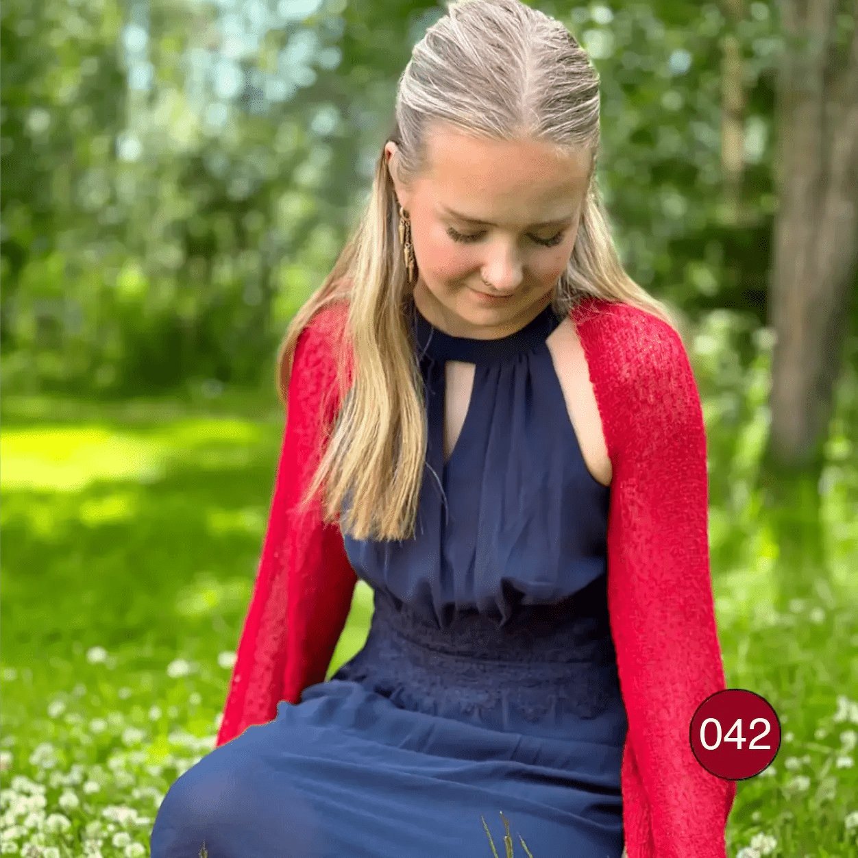 Ljushårig kvinna i en röd Liv bolero över en marinblå klänning, med en tunn halsduk knuten om halsen; den korta finstickade boleron sitter tätt över axlarna och värmer armarna med långa ärmar. Ulla Jacobsson Kläder