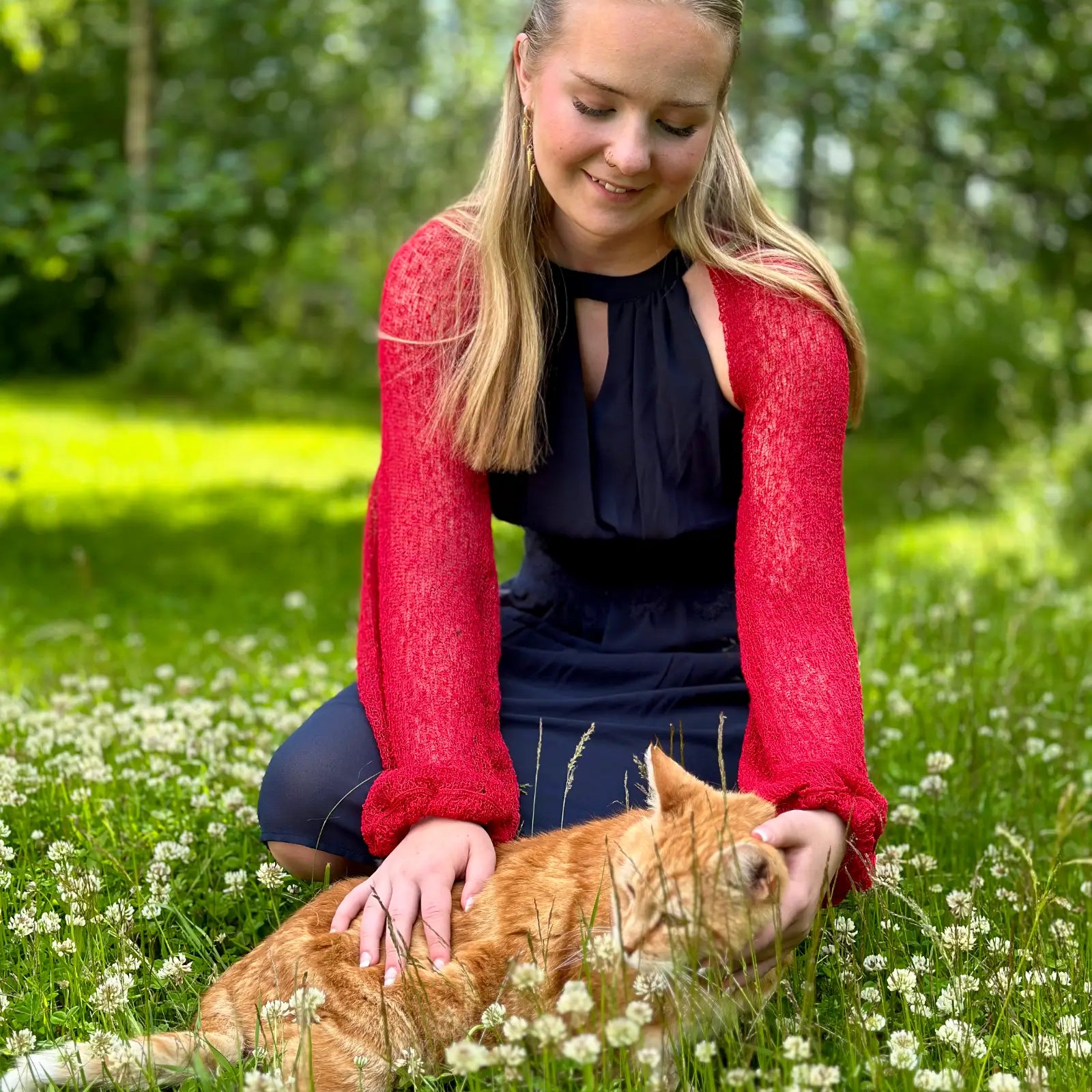 Ljushårig kvinna sitter i en sommaräng med en orange katt i famnen, klädd i en röd Liv bolero över en marinblå klänning; den korta finstickade boleron värmer axlarna mot det höga gräset. Ulla Jacobsson Kläder