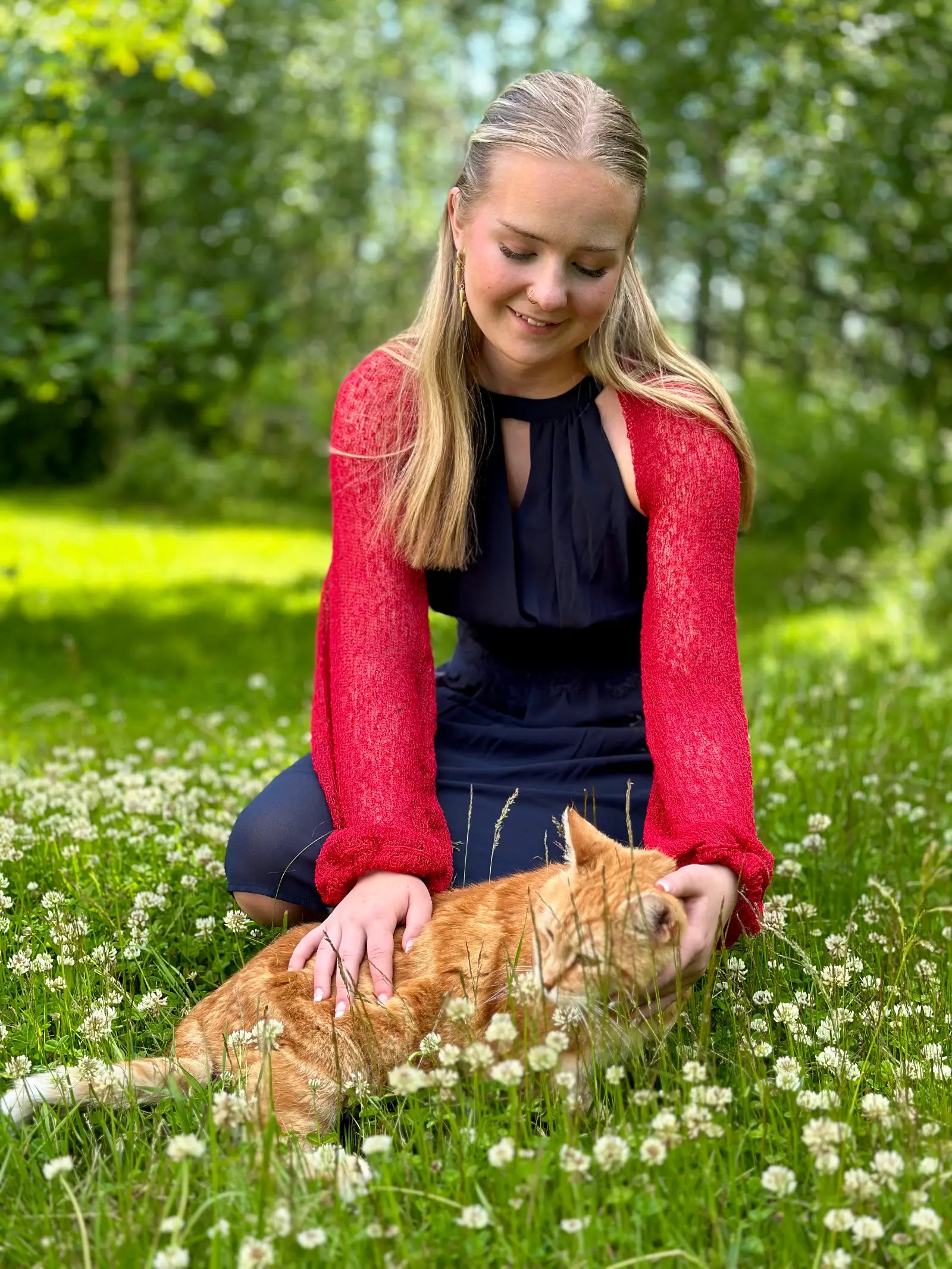 Ljushårig kvinna sitter i en sommaräng med en orange katt i famnen, klädd i en röd Liv bolero över en marinblå klänning; den korta finstickade boleron värmer axlarna mot det höga gräset. Ulla Jacobsson Kläder