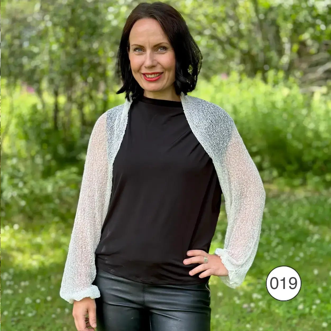 Mörkhårig kvinna på en grön sommaräng bär en vit Liv bolero öppet över ett svart linne; den finstickade boleron med långa ärmar ramar in axlarna och ger en lätt kontrast mot det mörka plagget. Ulla Jacobsson Kläder