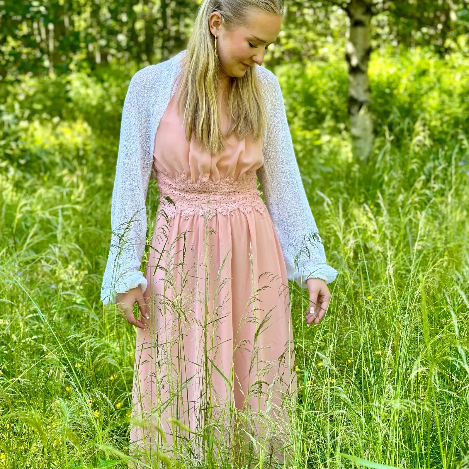 Ljushårig kvinna på en äng tittar ned i en vit Liv bolero buren över en aprikosfärgad klänning; det tunna finstickade garnet ligger mjukt över axlarna och ger ett ljust, lätt lager. Ulla Jacobsson Kläder