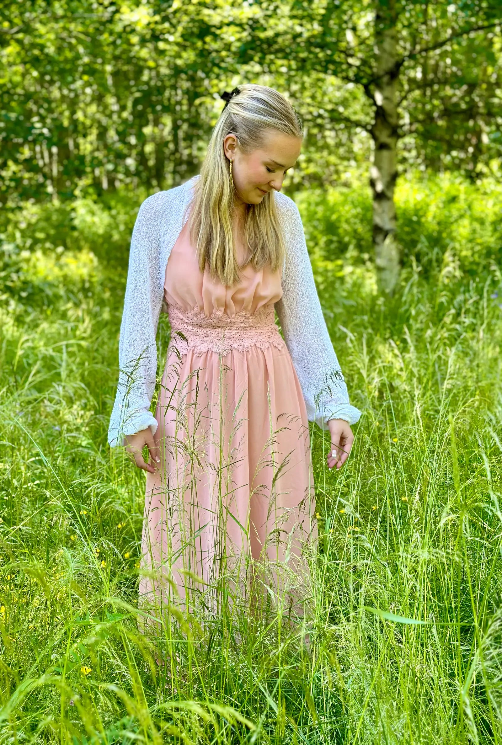 Ljushårig kvinna på en äng tittar ned i en vit Liv bolero buren över en aprikosfärgad klänning; det tunna finstickade garnet ligger mjukt över axlarna och ger ett ljust, lätt lager. Ulla Jacobsson Kläder