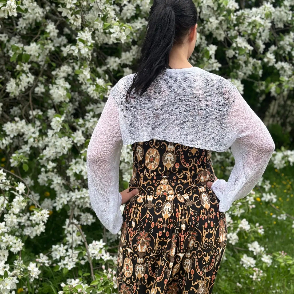 Mörkhårig kvinna fotograferad bakifrån vid blommande buskar i en vit Liv bolero ovanpå en mönstrad klänning; det luftiga finstickade garnet faller kort över ryggen och de långa ärmarna ramar in armarna. Ulla Jacobsson Kläder