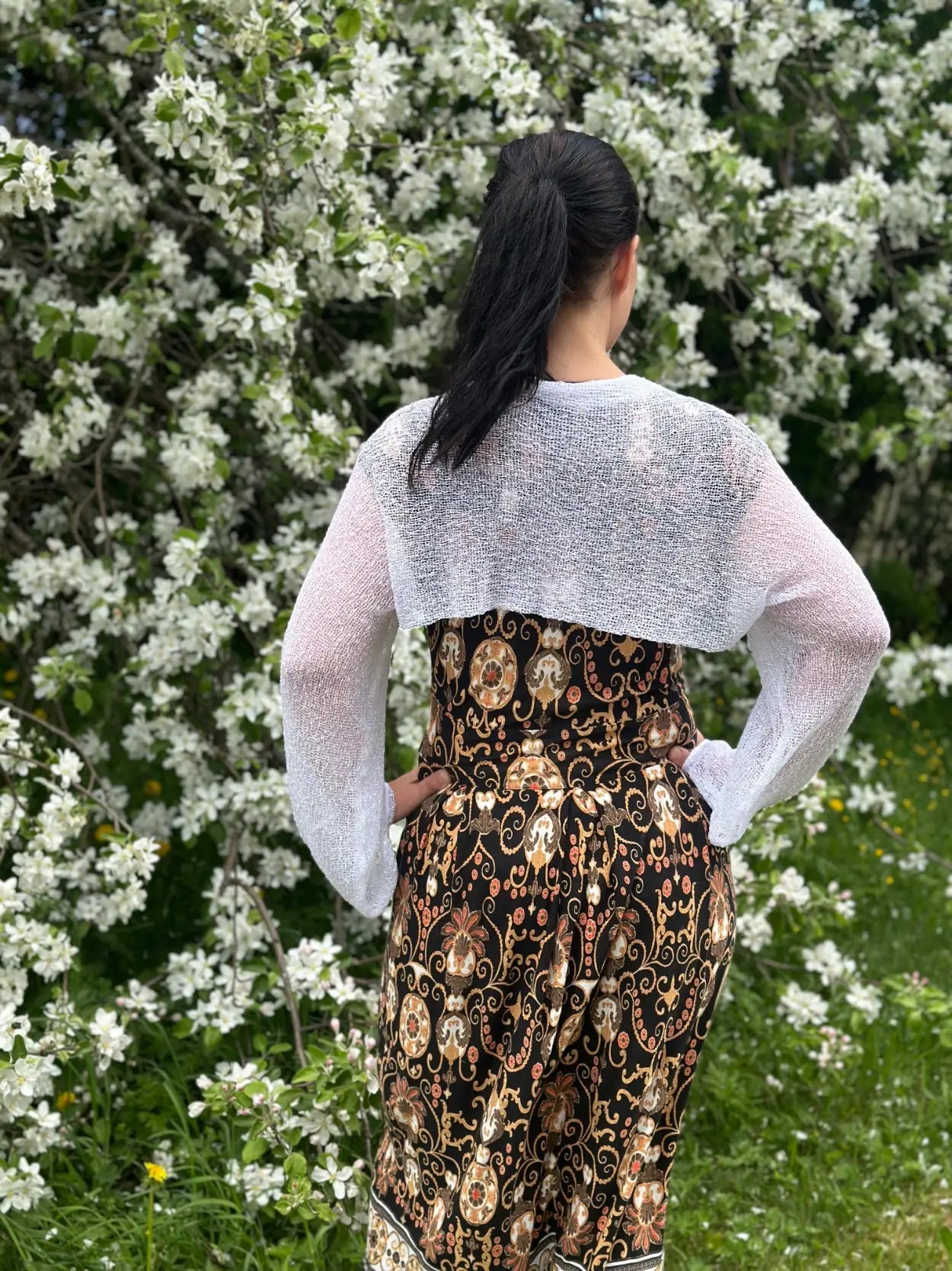 Mörkhårig kvinna fotograferad bakifrån vid blommande buskar i en vit Liv bolero ovanpå en mönstrad klänning; det luftiga finstickade garnet faller kort över ryggen och de långa ärmarna ramar in armarna. Ulla Jacobsson Kläder