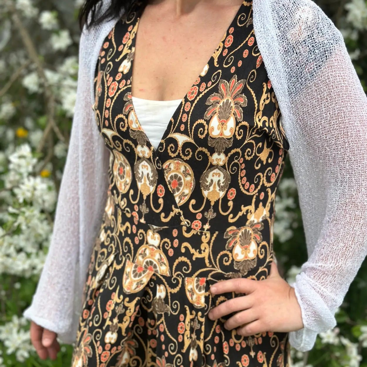 Mörkhårig kvinna vid blommande buskar i en vit Liv bolero buren öppet över en mörk mönstrad klänning; den tunna finstickade boleron med långa ärmar ger en elegant, ljus lagereffekt till den figursömmade klänningen. Ulla Jacobsson Kläder