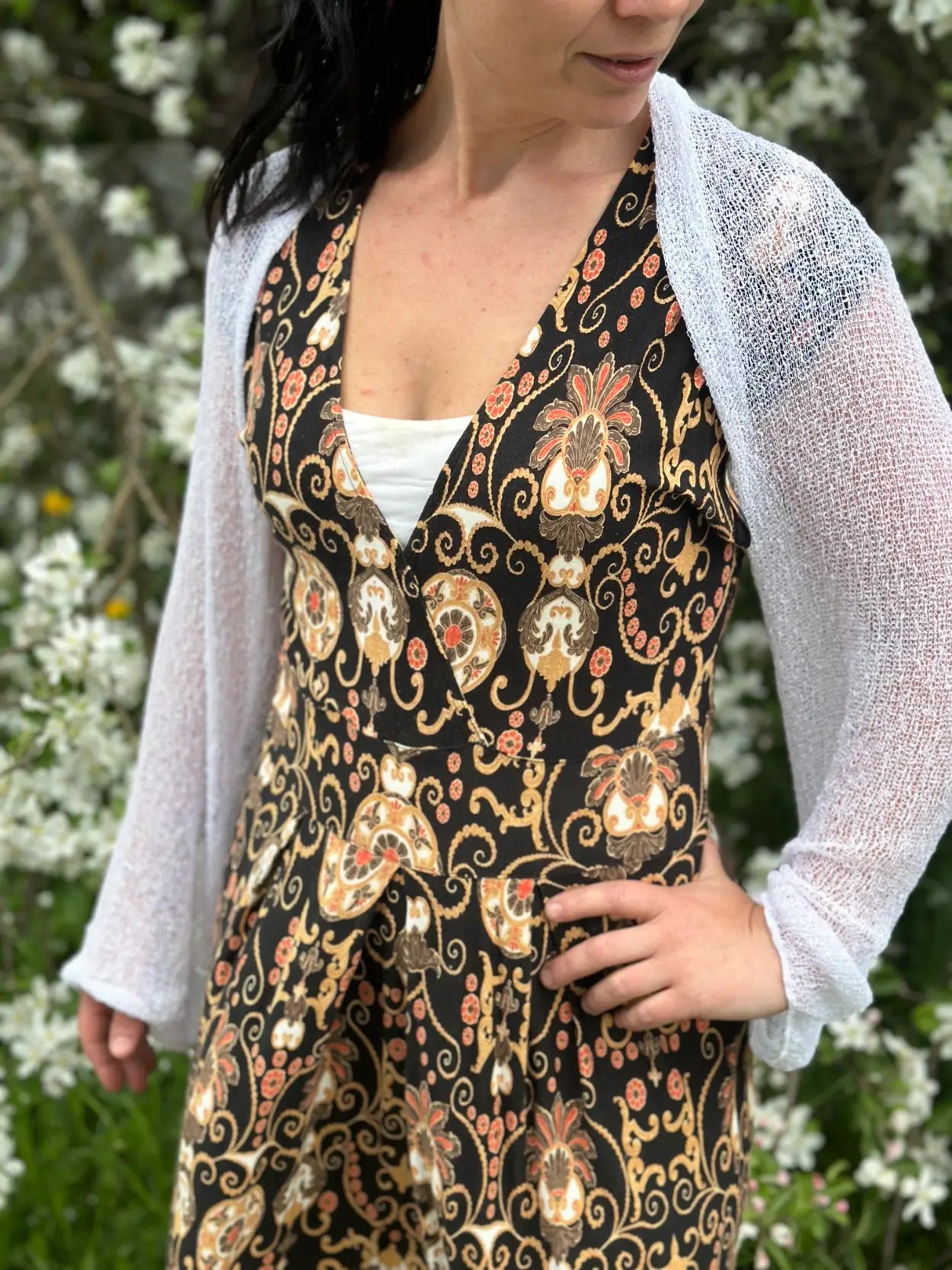 Mörkhårig kvinna vid blommande buskar i en vit Liv bolero buren öppet över en mörk mönstrad klänning; den tunna finstickade boleron med långa ärmar ger en elegant, ljus lagereffekt till den figursömmade klänningen. Ulla Jacobsson Kläder