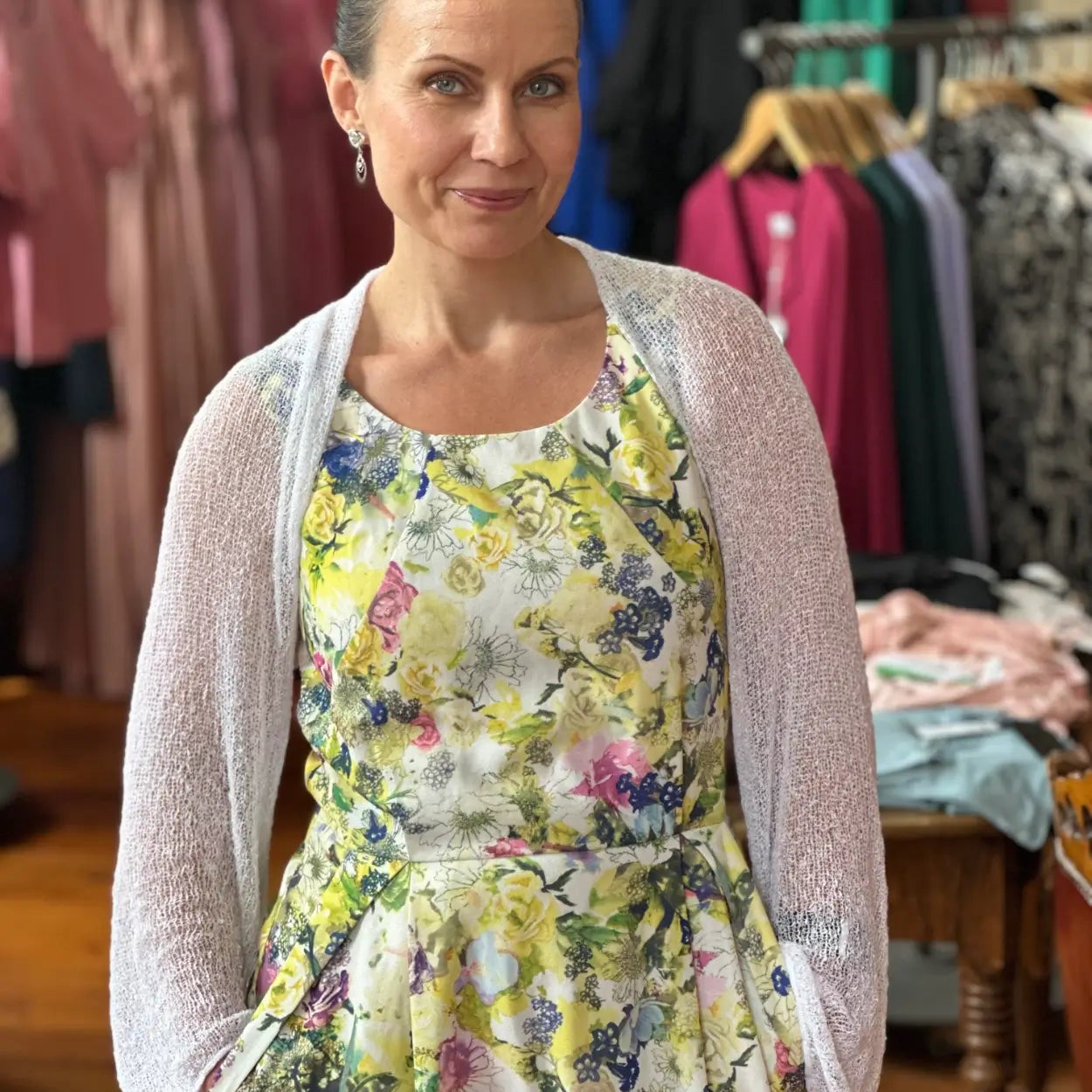 Mörkhårig kvinna inomhus i en vit Liv bolero ovanpå en gulblommig klänning; det tunna finstickade garnet ligger luftigt över axlarna och ger en lätt, festlig touch till sommarklänningen. Ulla Jacobsson Kläder