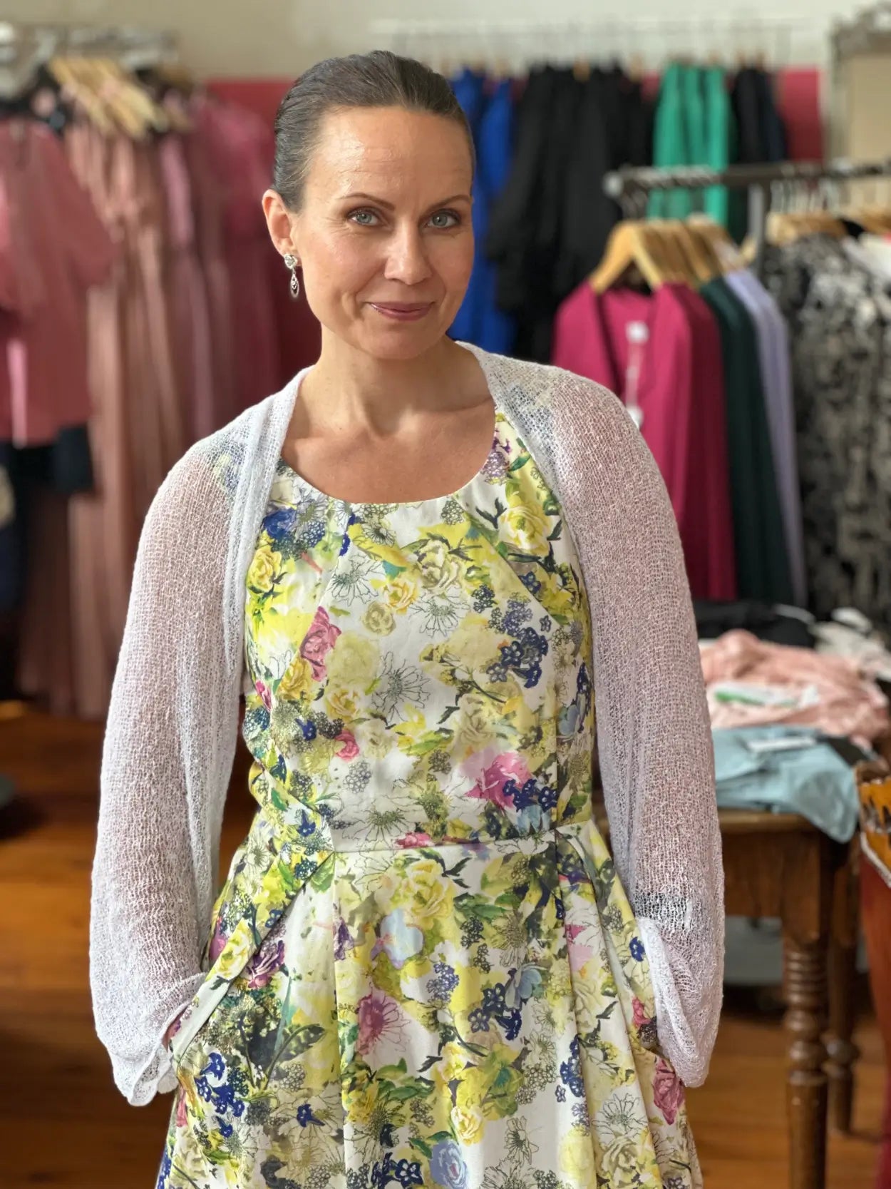 Mörkhårig kvinna inomhus i en vit Liv bolero ovanpå en gulblommig klänning; det tunna finstickade garnet ligger luftigt över axlarna och ger en lätt, festlig touch till sommarklänningen. Ulla Jacobsson Kläder