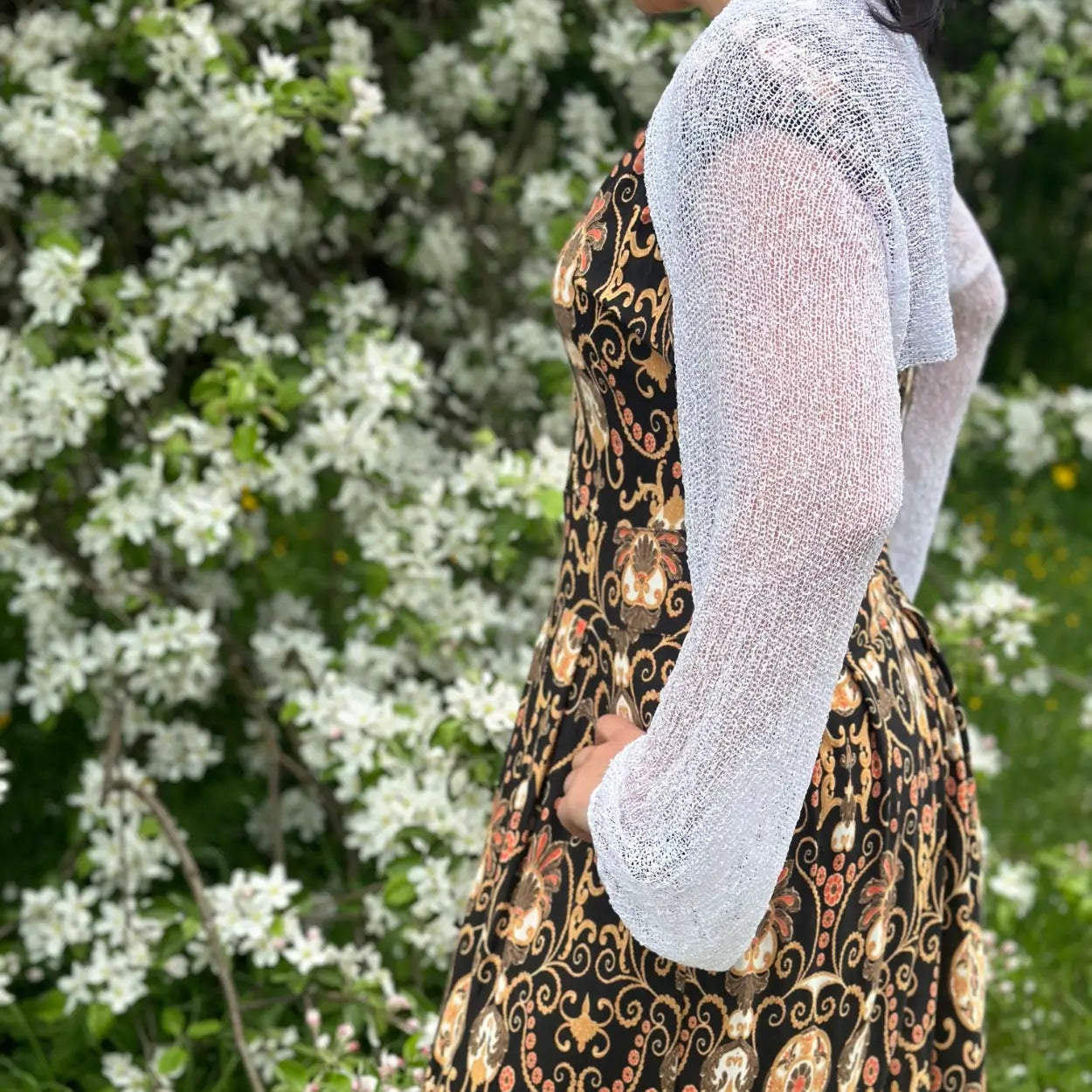 Mörkhårig kvinna snett bakifrån i en vit Liv bolero över en mönstrad klänning; den korta finstickade boleron värmer axlar och armar och låter mönstret skymta igenom det luftiga garnet. Ulla Jacobsson Kläder