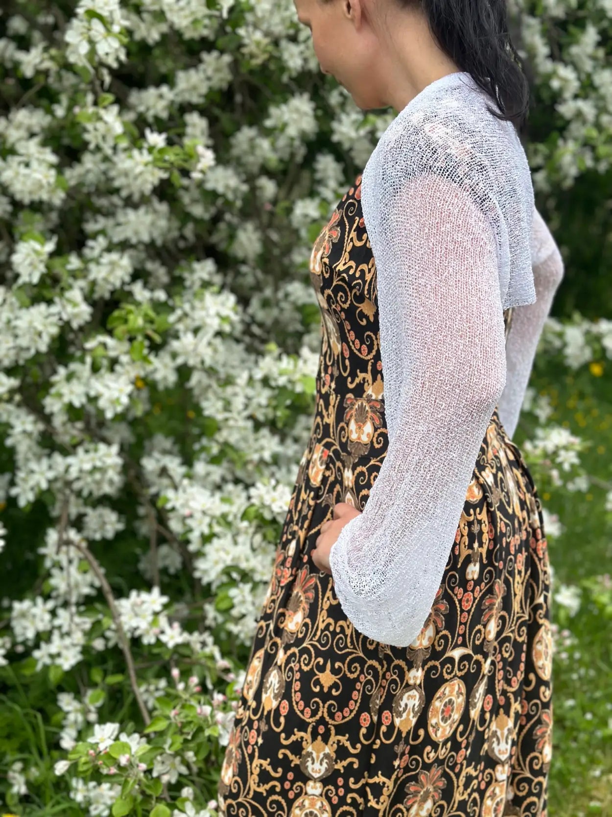 Mörkhårig kvinna snett bakifrån i en vit Liv bolero över en mönstrad klänning; den korta finstickade boleron värmer axlar och armar och låter mönstret skymta igenom det luftiga garnet. Ulla Jacobsson Kläder