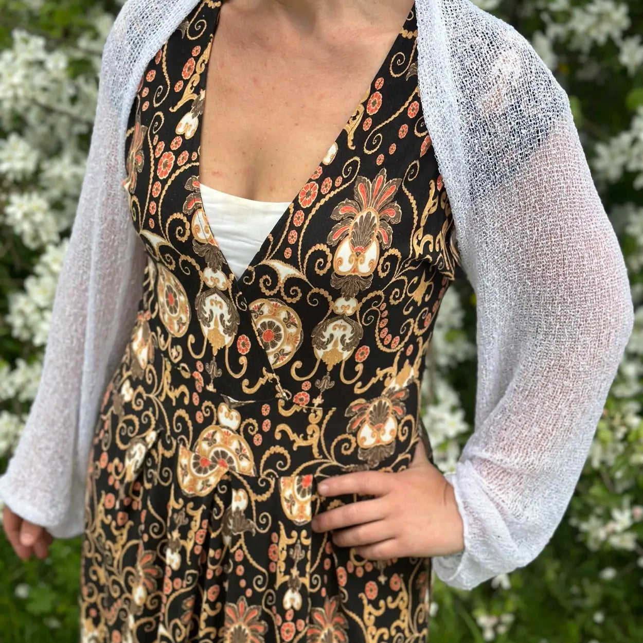 Mörkhårig kvinna i en vit Liv bolero buren öppet över en mörk mönstrad klänning; det tunna finstickade garnet skymtar nästan genomskinligt och lägger ett lätt, ljust lager över axlarna. Ulla Jacobsson Kläder