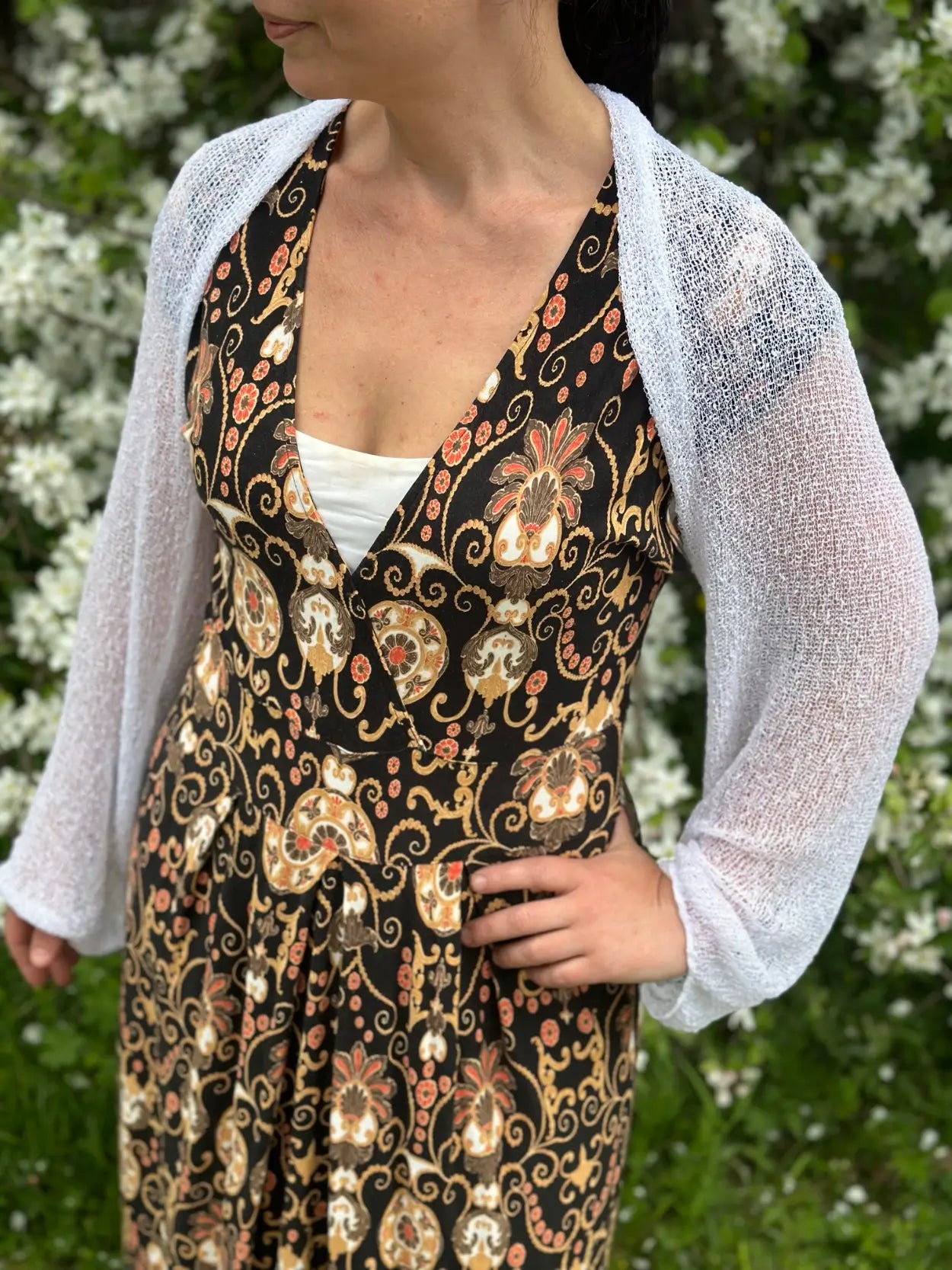 Mörkhårig kvinna i en vit Liv bolero buren öppet över en mörk mönstrad klänning; det tunna finstickade garnet skymtar nästan genomskinligt och lägger ett lätt, ljust lager över axlarna. Ulla Jacobsson Kläder