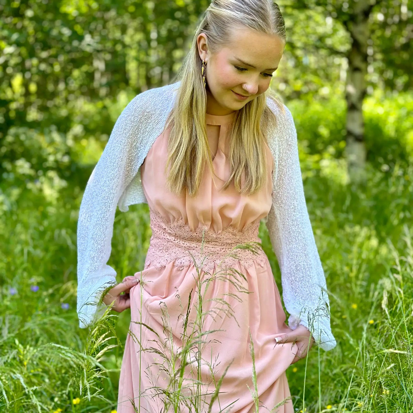 Ljushårig kvinna på en sommaräng i en vit Liv bolero ovanpå en pudrig rosa klänning; den finstickade boleron med långa ärmar värmer axlarna och ramar mjukt in den ljusa klänningen. Ulla Jacobsson Kläder