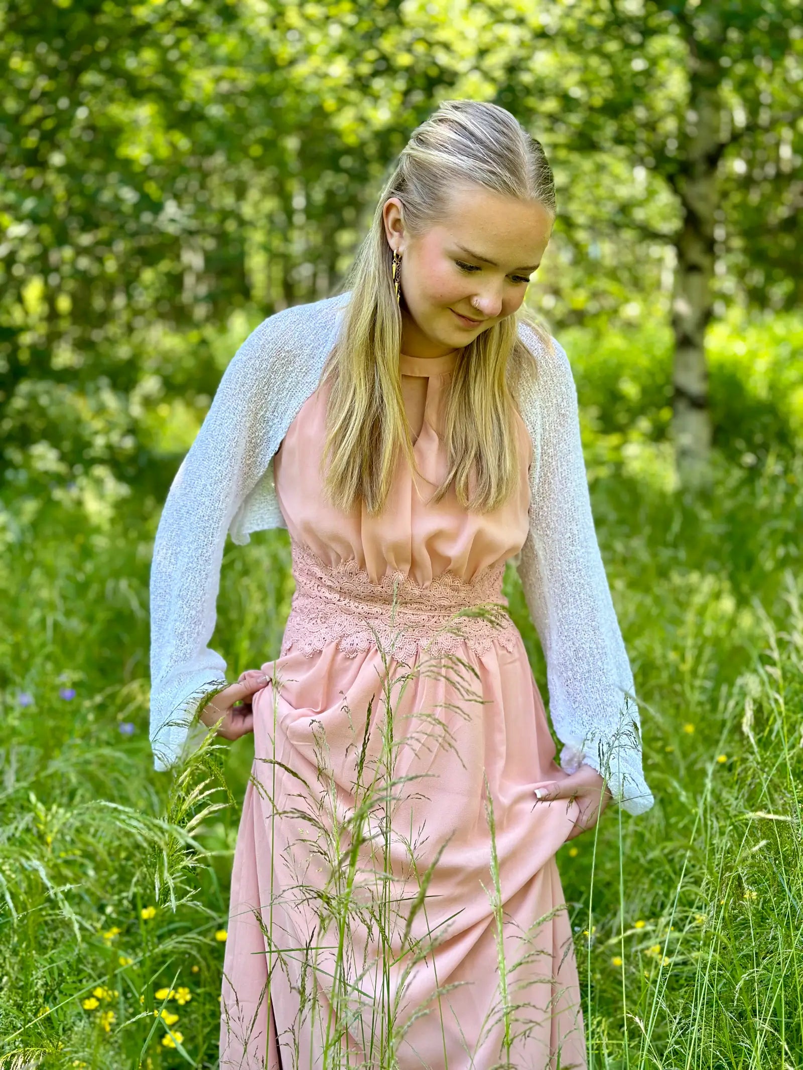 Ljushårig kvinna på en sommaräng i en vit Liv bolero ovanpå en pudrig rosa klänning; den finstickade boleron med långa ärmar värmer axlarna och ramar mjukt in den ljusa klänningen. Ulla Jacobsson Kläder
