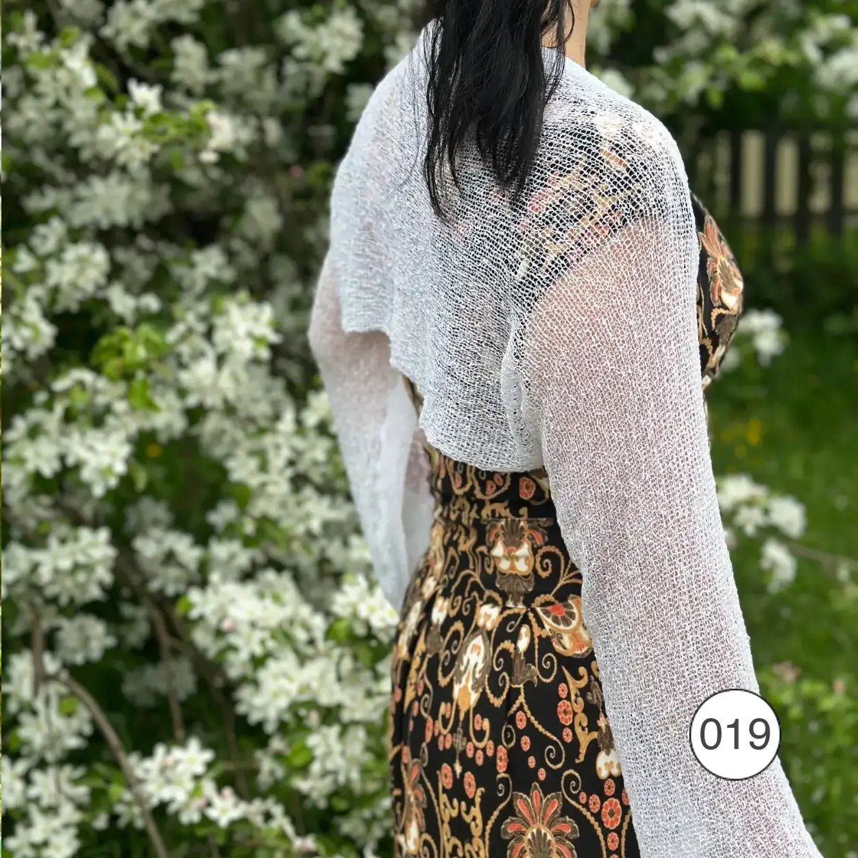 Mörkhårig kvinna vid blommande buskar, sedd snett bakifrån i en vit Liv bolero över en mönstrad klänning; det luftiga finstickade garnet faller kort över ryggen och de långa ärmarna ramar in armarna. Ulla Jacobsson Kläder