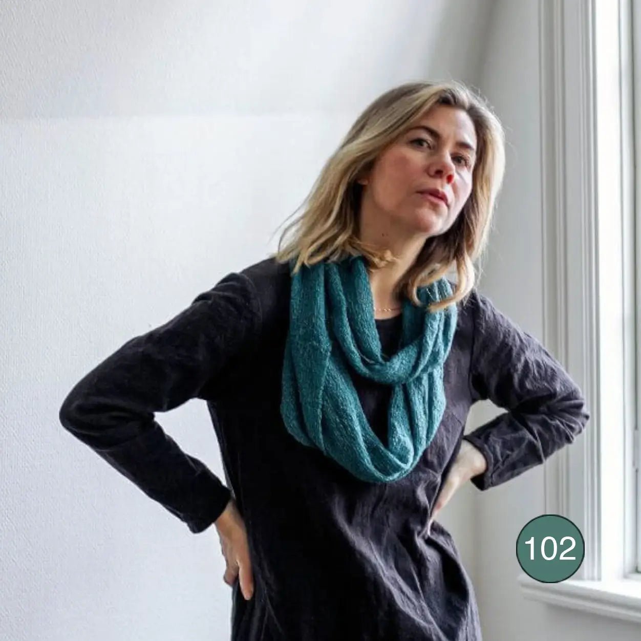 Blond kvinna vid ett fönster i svart topp, handen i sidan, med en buteljgrön Sally tubsjal virad runt halsen; det finstickade garnet i djup grönton faller mjukt. Ulla Jacobsson Kläder