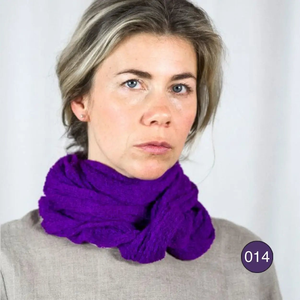 Blond kvinna mot ljus bakgrund med en mörklila Sally tubsjal virad runt halsen; det tunna, finstickade garnet lägger sig mjukt i flera lager mot hakan. Ulla Jacobsson Kläder