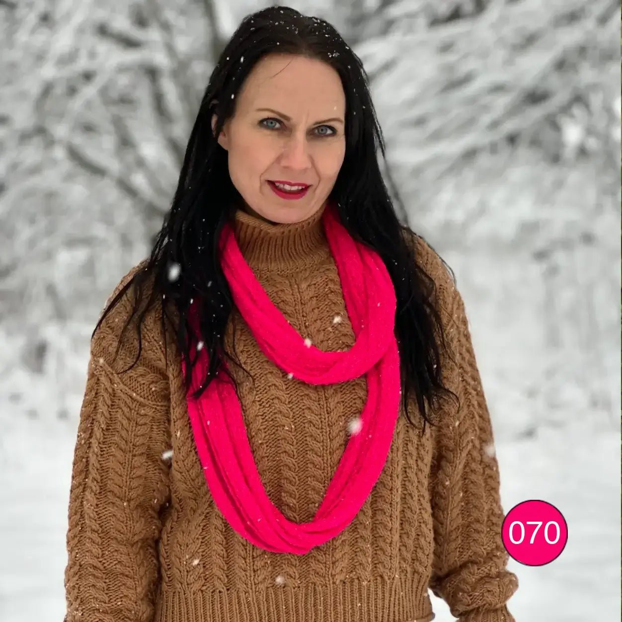 Mörkhårig kvinna i kameltröja med en neonrosa Sally tubsjal hängande i en lång, lös ögla längs överkroppen; det tunna finstickade garnet kan viras eller bäras löst. Ulla Jacobsson Kläder