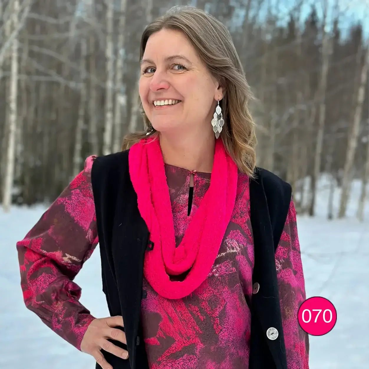 Kvinna i mönstrad topp i björkskog med en neonrosa Sally tubsjal virad runt halsen; det finstickade garnet lyser klart cerise och faller mjukt i flera varv. Ulla Jacobsson Kläder