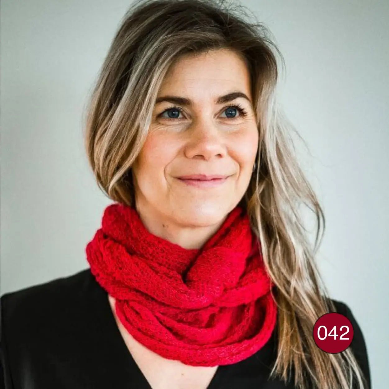 Mörkblond kvinna i närbild mot neutral bakgrund, med en röd Sally tubsjal virad tätt runt halsen; det finstickade garnet är tunt och lätt men fylligt i flera lager. Ulla Jacobsson Kläder