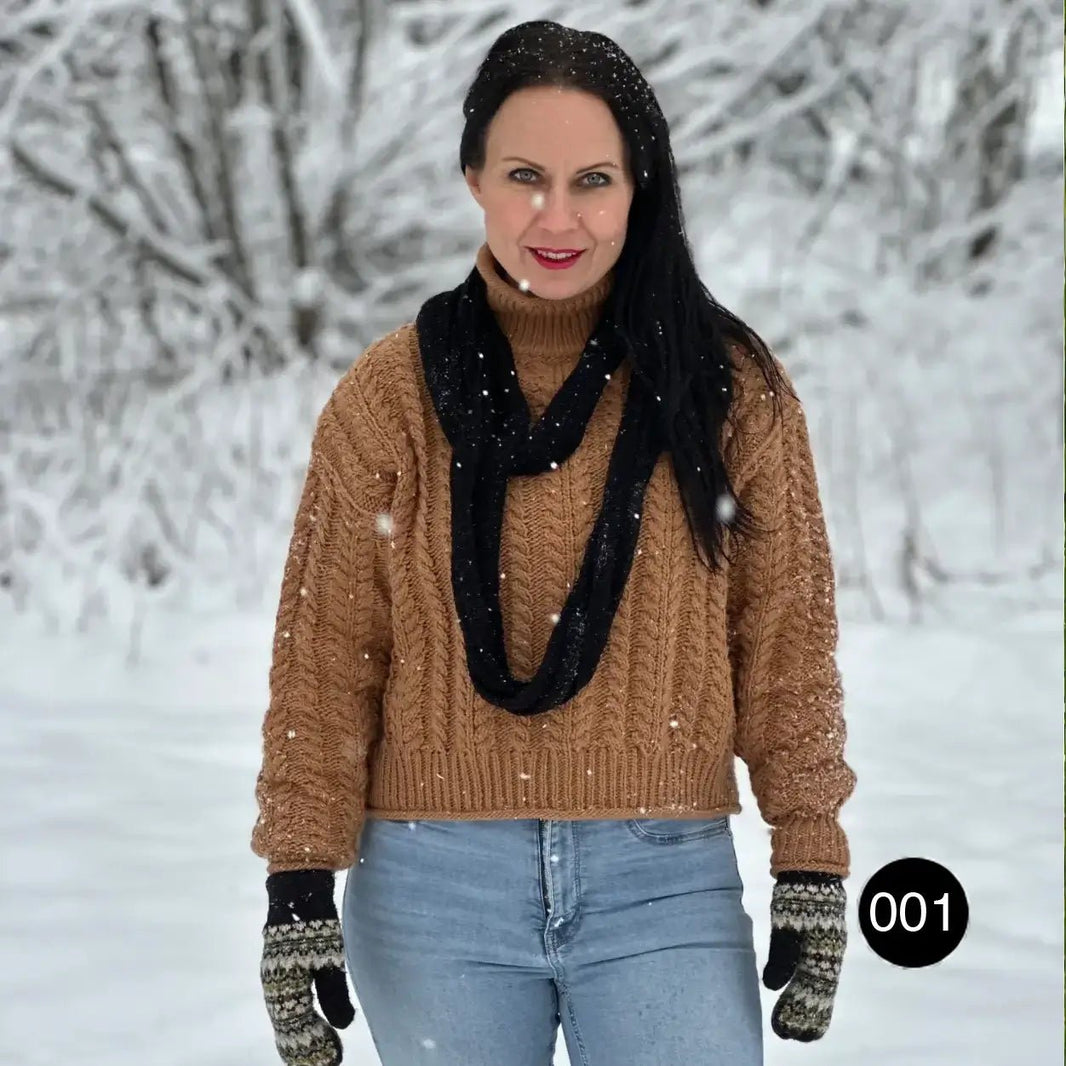 Mörkhårig kvinna i snön i jeans och handskar, med en svart Sally tubsjal hängande i en lång, lös ögla längs överkroppen; det tunna finstickade garnet kan viras tätt eller bäras löst. Ulla Jacobsson Kläder