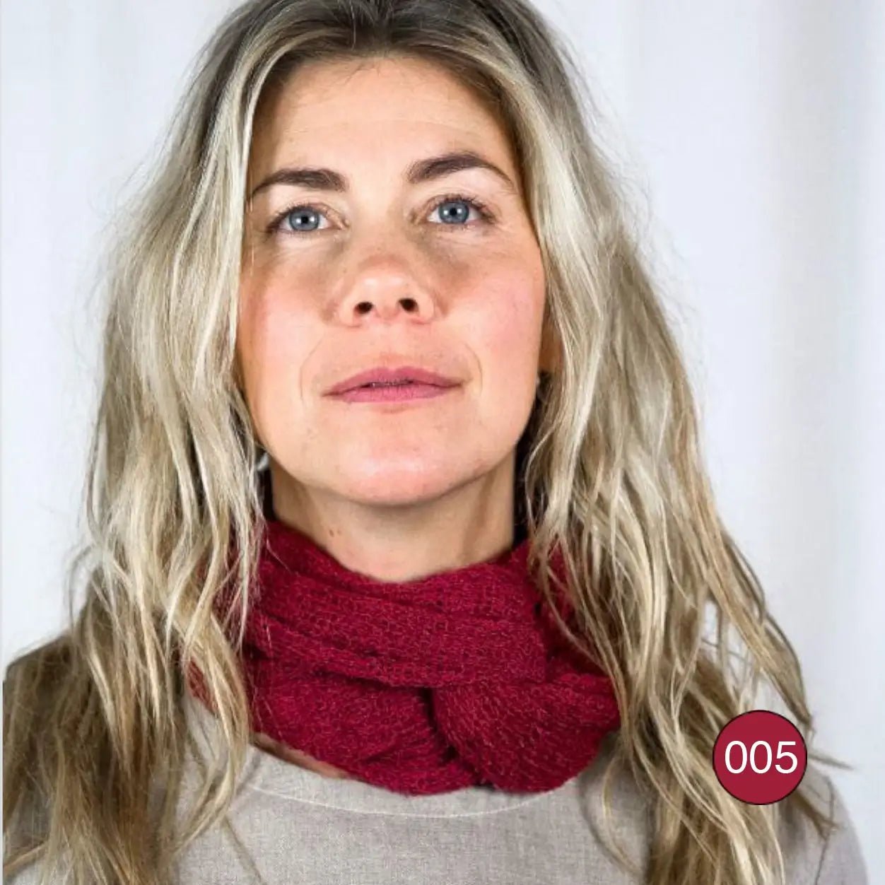 Blond kvinna tittar uppåt i grå tröja med en vinröd Sally tubsjal runt halsen; det finstickade garnet i djup vinröd ton ligger luftigt och omsvepande. Ulla Jacobsson Kläder